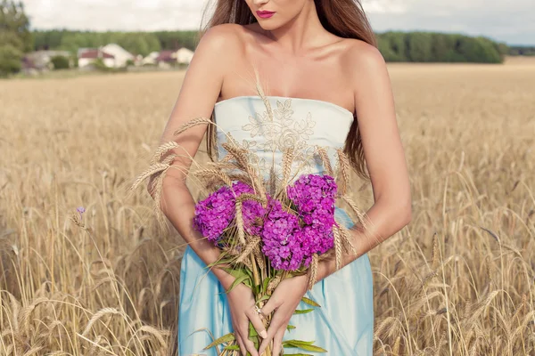Красивая сексуальная худенькая девушка в голубом платье в поле с букетом цветов и колосьев в его руках на закате в Солнечный день — стоковое фото