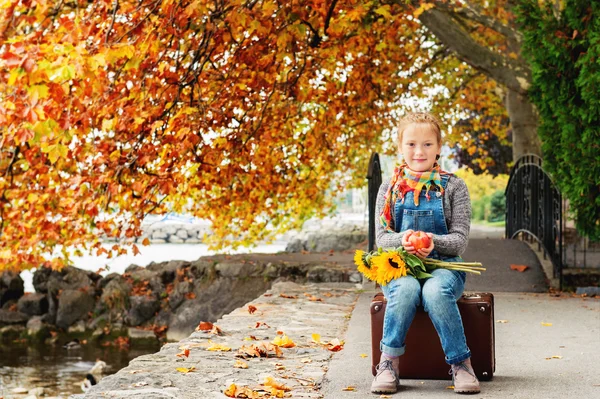 Осенний портрет очаровательны блондинка девочка 8 лет, носить теплые пуловеры, джинсовый комбинезон и бежевые туфли, сидя на старый старинных чемодан, красное яблоко и Букет желтых подсолнухов — стоковое фото
