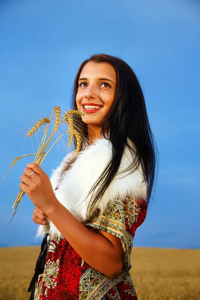 Молодая женщина с орнаментальный платье и белый мех, стоящий на пшеничном поле с захода солнца. Холдинг букет — стоковое фото